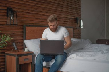 Yakışıklı bir adam elinde bir bilgisayarla yatağın kenarında oturur.
