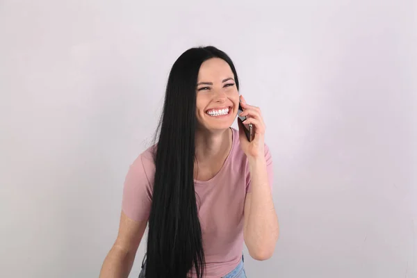 Молода приваблива брюнетка з довгим волоссям, що стоїть на сірому фоні в рожевій сорочці, має телефонну розмову і сміється отримувати хороші новини і бути дуже щасливим — стокове фото