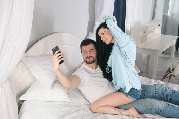 Jovem casal bonito e amoroso tirar foto selfie na câmera do smartphone enquanto sentado na cama na manhã — Fotografia de Stock