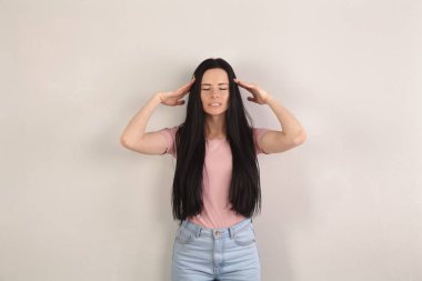 Çekici genç esmer kadın uzun saçlı having güçlü baş ağrısı ve baş tarafından el ele tutuşarak gri arka plan tarafından duruyor. Kot pantolon ve pembe gömlek giyiyor