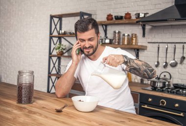 Genç mutlu yakışıklı gülümseyen adam mutfakta kahvaltı hazırlamak süt ile tahıl karıştırma ve telefon görüşmesi var