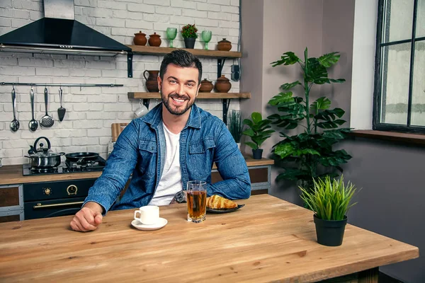 Handsome young smiling guy is home at his kitchen eating breakfast. croissant, coffee and an apple juice — Stock Photo, Image