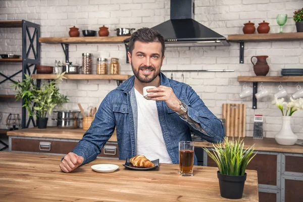 Young handsome smiling man is drinking his morning coffee with croissant and an apple juice at the kitchen — Stock Photo, Image