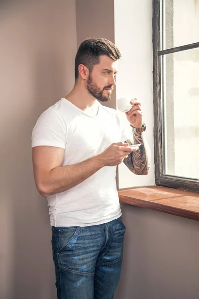 young handsome thoughtful man is looking at the window and having his morning coffee