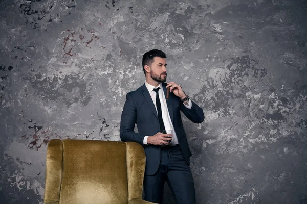 Un hombre de negocios guapo y guapo está descansando en la oficina parado junto a la silla y bebiendo whisky con un cigarro y pensando. llevar traje y corbata — Foto de Stock