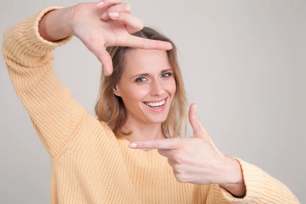 Portrait of young blonde woman with cheerful happy expression on her face, with good mood, making square with her fingers. wearing yellow sweater. poses against grey background. face expression, emoti — Stock Photo, Image