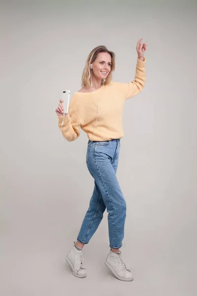 Знімок молодої блондинки танцює, тримаючи свій смартфон в руці і слухаючи музику в навушниках і посміхаючись. йди і танцюй зі мною. в жовтому светрі і блакитних джинсах. пози — стокове фото