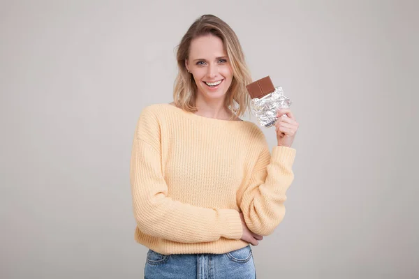 Ein schönes junges zartes Mädchen mit blonden Haaren hält eine Schokoriegel ins Gesicht, die sie in einem gelben Pullover trägt. posiert vor weißem Hintergrund. Mimik, Gefühl. — Stockfoto