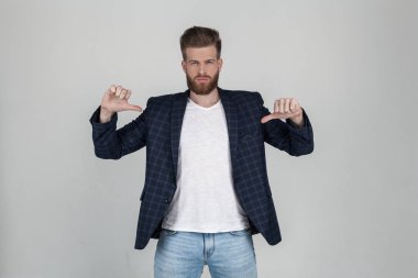 Güzel bir saç modeli ile güzel bir seksi sakallı adam onun t-shirt parmaklarını işaret. metin veya logo için yer. bir ceket giymiş, beyaz arka plan önünde pozlar.