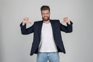 Güzel bir saç modeli ile gülümseyen seksi sakallı adam onun t-shirt parmaklarını işaret ediyor. metin veya logo için yer. bir ceket giymiş, beyaz arka plan önünde pozlar.
