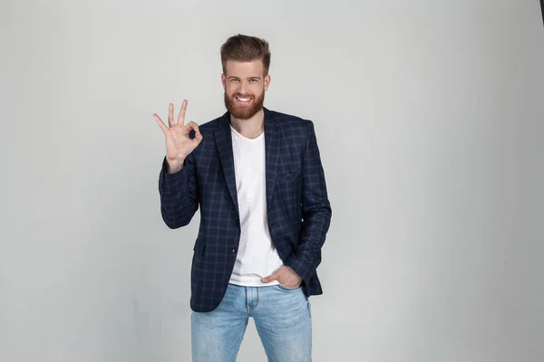 Apuesto negocio sonriente mostrando gesto bien. vestido con chaqueta. posa sobre fondo gris. expresión facial, emociones . — Foto de Stock