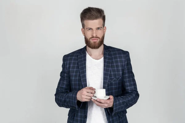 Un hermoso hombre de negocios barbudo sexy con una chaqueta mira a la cámara. sosteniendo una taza de café en sus manos se para frente a un fondo blanco — Foto de Stock