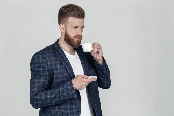 En sexy, skjeggete forretningsmann i jakke ser på det ytterste i profilen. Han holder en kopp kaffe i hendene og står foran en hvit bakgrunn. – stockfoto