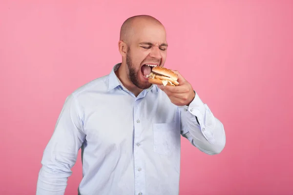 A satisfied man with a lush beard and a bald head. opens a very wide mouth so as soon as possible to eat his burger. stand in front of the pink background