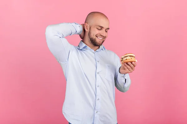 A satisfied man with a lush beard and a bald head. looks at his burger with a thirst for him to eat it as soon as possible. stand in front of the pink background