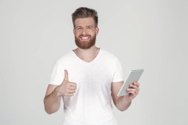 bir sakal ve rahat giyim giymiş şık bir saç modeli ile güzel bir adam. gülümseyerek üst bir başparmak gösterir ve kameraya bakar, tablet tutar . beyaz arka planın karşısında duruyor