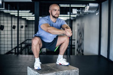 Spor salonundaki bir çömelme kutusunda çömelmiş bir adam. spor salonunda ki adam tahta bir çömelmiş kutunun üzerinde çalışıyor. bir spor üniforması giymiş.