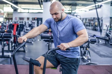 Spor salonunda egzersiz bisikleti kullanan genç bir sporcu. Spor salonunda spor yapmak ve müzik dinlemek için hava bisikleti kullanan spor adamı. Spor kıyafetleri içinde.