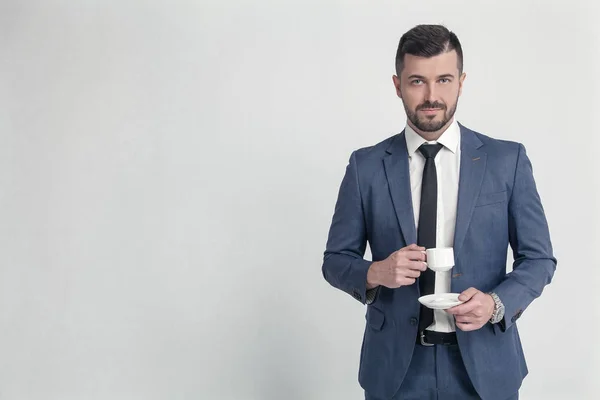 Stylish bearded businessman holding coffee cup and looking at camera isolated on grey background on the left space for your logo or text