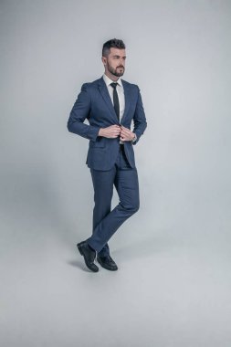 çarpıcı, şık, serin, erkeksi, mavi takım elbiseli sakallı zengin adam iki kollu ceket üzerinde düğme yitirme, yan bakarak, gri arka plan üzerinde izole tam uzunlukta portre