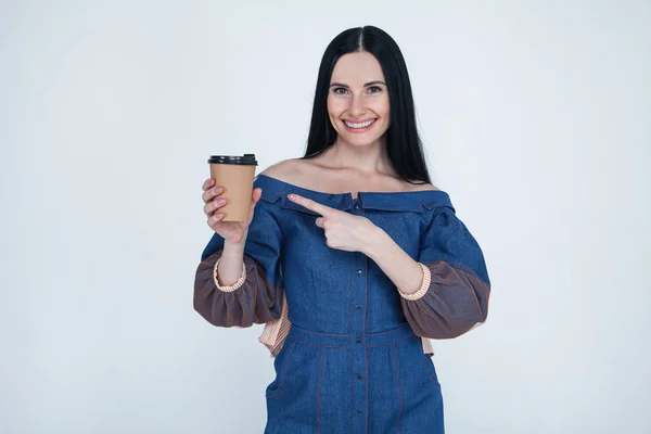 Porträt von attraktiven hübsch schönen stilvollen netten fröhlichen brünetten Mädchen in Jeans Kleid, zeigt auf Pappbecher Kaffee, isoliert auf grauem Hintergrund — Stockfoto