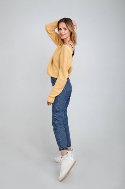 İnce ve güzel. Gri arka plan aleyhine dururken rahat giyim gülümseyen ve elinden saç tutarak çekici genç kadının tam uzunlukta stüdyo çekim