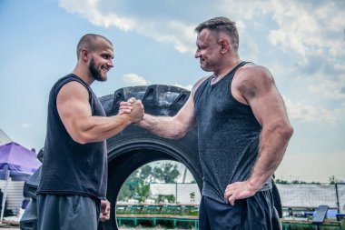 Merhaba sporcular. İki atlet sokak spor salonunda el sıkışıyor. İki güzel adam büyük lastiğin arasında duruyor. Kameratam uzunlukta emin iki Güçlü adam
