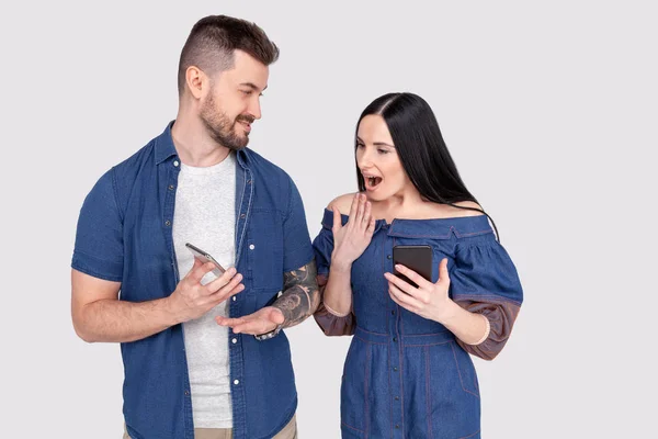 Jeune homme non rasé en jeans chemise tenant smartphone montrant quelque chose sur téléphone portable à sa femme. Surpris jeune femme regardant avec les yeux pleins d'incrédulité dans le smartphone de son mari — Photo