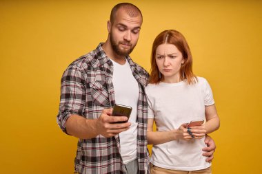 Cep telefonu ile ayakta genç bir çift portresi, adam sarı arka plan üzerinde izole yakın duran kızgın kız cep telefonu kullanıyor