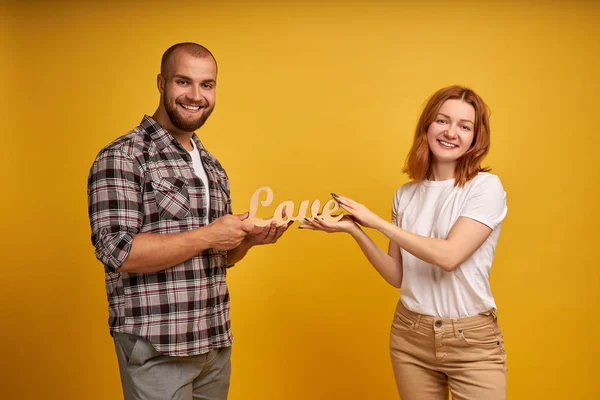 Portrait de charmant homme et femme charismatique tenant une statuette en bois du mot Amour. isolé sur fond jaune — Photo