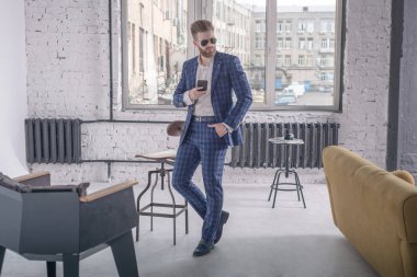 Mükemmel görünüyorsun. Evde dururken tam takım elbise ve güneş gözlüğü yakışıklı genç sakallı adam tam uzunlukta uzağa bakıyor. Cep telefonunu kullanarak.