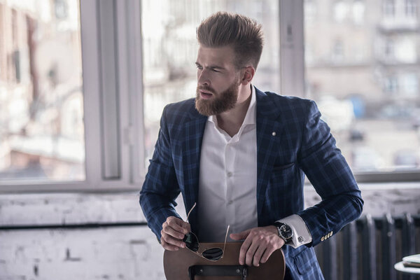 Amazing look. Good looking young bearded man in full suit looking away while sitting on the stool