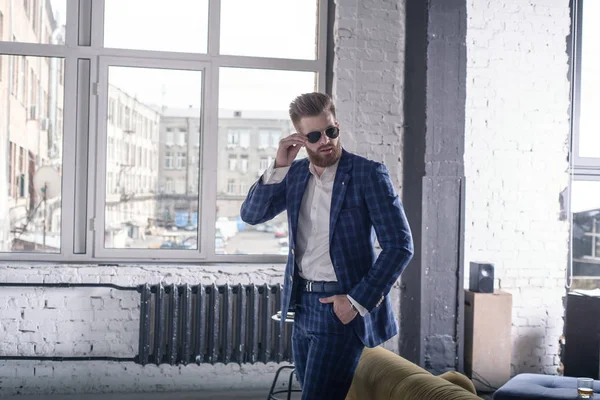 Es hora de irse. Longitud completa de joven guapo en traje completo y tirantes mirando hacia otro lado y manteniendo gafas de sol por su mano mientras está de pie en el interior del loft —  Fotos de Stock