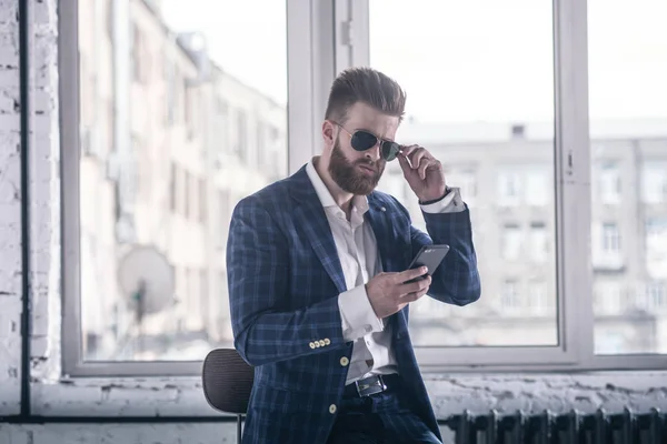 Él derretirá tu corazón. Hombre barbudo joven guapo en traje completo y gafas mirando a su teléfono celular mientras está sentado en el taburete — Foto de Stock