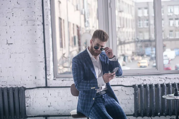 Él derretirá tu corazón. Hombre barbudo joven guapo en traje completo y gafas mirando a su teléfono celular mientras está sentado en el taburete —  Fotos de Stock
