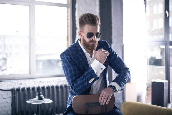 Un pensador guapo. Hombre joven de buen aspecto en traje completo y gafas manteniendo las manos apretadas mientras está sentado en el taburete — Foto de Stock