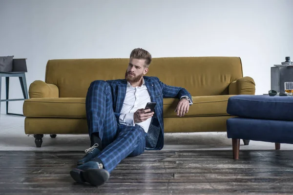 Te ves perfecta. Hombre joven guapo en traje completo usando su teléfono celular mientras está sentado en el suelo en casa —  Fotos de Stock