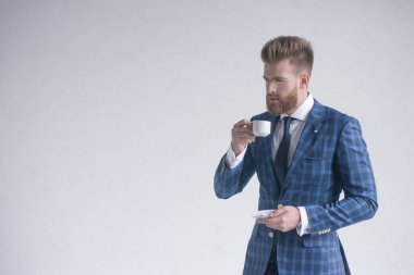 Kahve molası. Kahve fincanı tutan ve gri arka plan önünde dururken uzağa bakan tam takım elbiseli kendine güvenen genç adam. Logonuz veya metniniz için boş alan