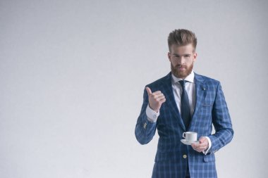 Kahve molası. Kahve fincanı tutan ve gri arka plan önünde dururken kameraya bakan tam takım elbiseli kendinden emin genç adam. Logonuz veya metniniz için boş alan