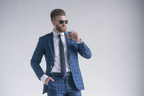 Pohledný muž drží sklenici whisky a dívá se stranou, když stojí před šedým pozadím. Volné místo pro logo nebo text. — Stock fotografie