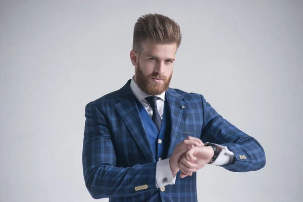 Joven barbudo elegante hombre de negocios líder en el interior vestido con traje de tres piezas comprobar la hora en su reloj. aislado en gris —  Fotos de Stock