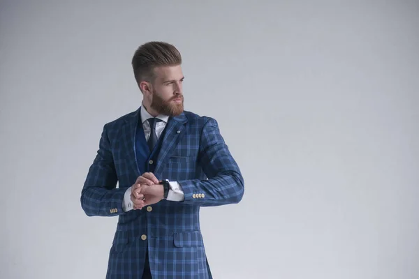 Young bearded stylish businessman leader indoors wearing three-piece suit with watch looking aside serious. isolated on grey. Place for a logo, advertisement or text — Stock Photo, Image