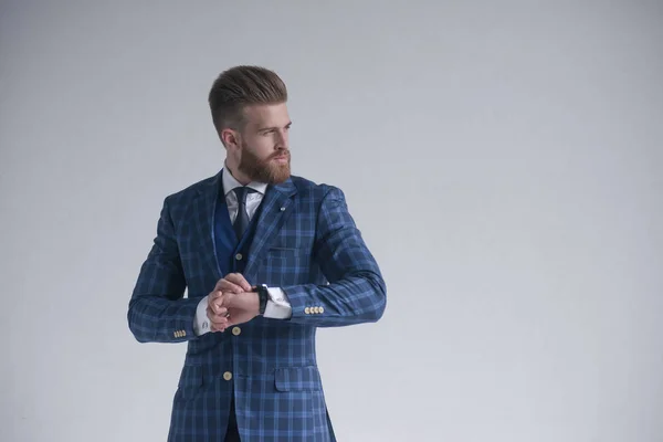 Joven barbudo elegante hombre de negocios líder en interiores con traje de tres piezas con reloj mirando a un lado serio. aislado en gris. Lugar para un logotipo, anuncio o texto — Foto de Stock