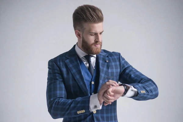 Joven barbudo elegante hombre de negocios líder en el interior vestido con traje de tres piezas comprobar la hora en su reloj. aislado en gris —  Fotos de Stock
