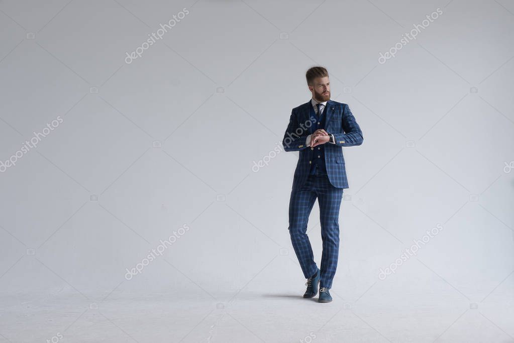 Young bearded stylish businessman leader indoors dressed in three-piece suit checking the time on his watch. isolated on grey.