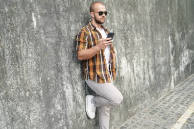 Genç iyi görünümlü cesur sakallı adam elinde cep telefonu ile gri modern Loft duvara karşı ayakta sohbet ve kulaklıklar müzik dinler. sarı gömlek, kot pantolon ve güneş gözlüğü giyiyor
