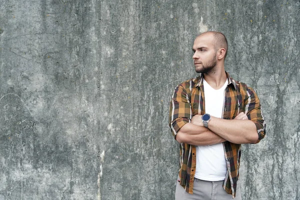 portrait of young good looking bold bearded guy standing outdoors against grey loft wall looking aside with his hands crossed.wearing yellow shirt and jeans. on the left place for your logo or text