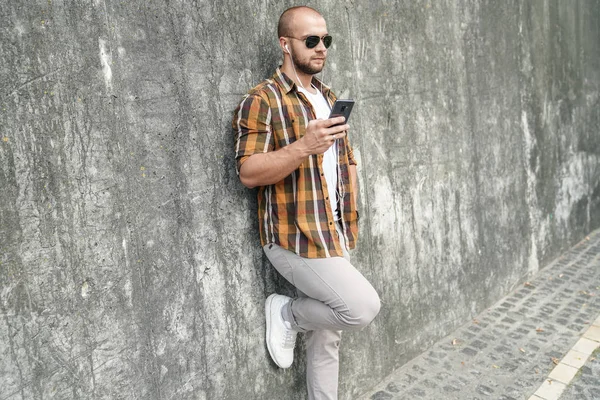 Genç iyi görünümlü cesur sakallı adam elinde cep telefonu ile gri modern Loft duvara karşı ayakta sohbet ve kulaklıklar müzik dinler. sarı gömlek, kot pantolon ve güneş gözlüğü giyiyor