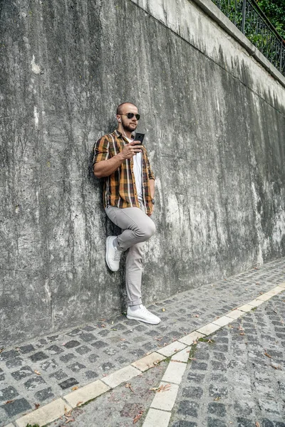 vertical shot of good looking bold bearded guy standing outdoors against grey wall with cell phone in his hand chatting and listens music in headphones. wearing yellow shirt, jeans and sunglasses.