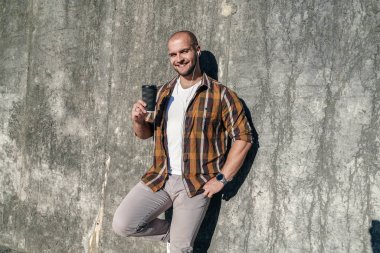 Genç iyi görünümlü kalın sakallı Freelancer ayakta açık havada gri modern Loft duvara tutarak kahve elinde ve kulaklıklar ve gülümseyen. sarı gömlek ve kot giyen.